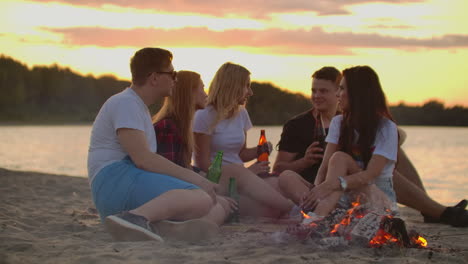 The-best-friends-from-university-are-sitting-in-around-bonfire-on-the-lake-coast.-They-are-talking-to-each-other-and-drinking-beer-at-sunset.